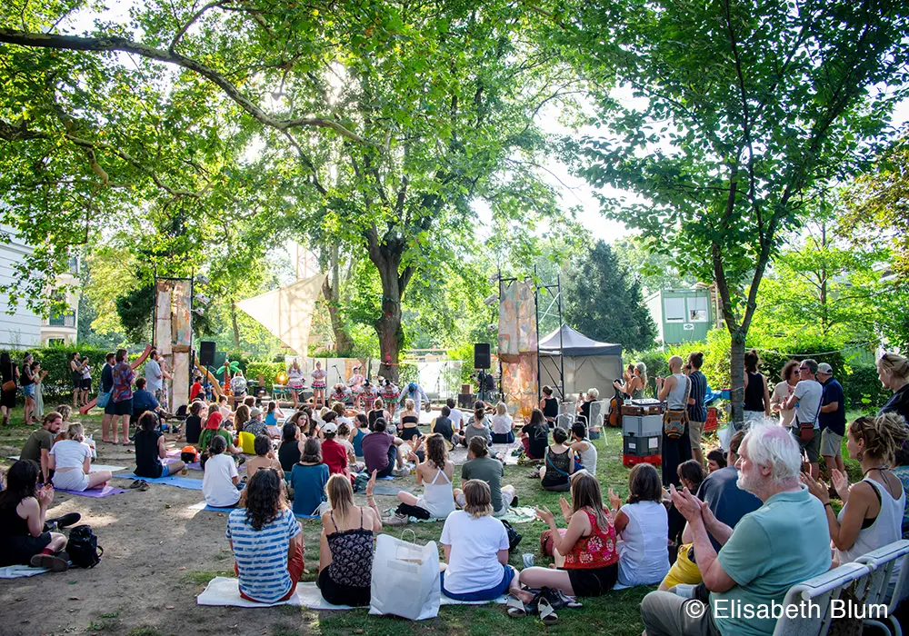 Crowd sitting on the lawn under trees at the event listening to musicians playing on a stage in the background.