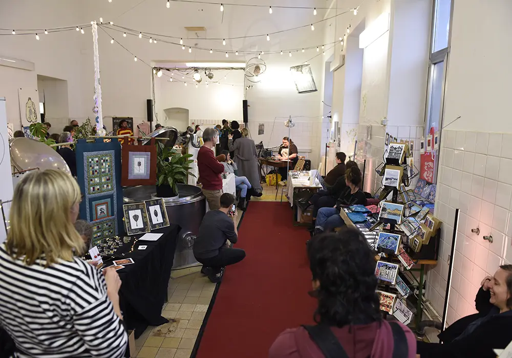 Market stall in a brightly lit room with several visitors. There are tables with handicrafts, plants and a cosy atmosphere. In the background a table with someone sitting behind